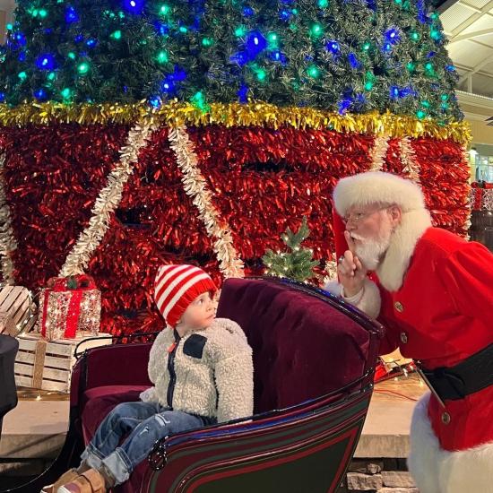 Santa is holding up his hand to his mouth saying "Shh" while sneaking up behind a toddler sitting in a sleigh.