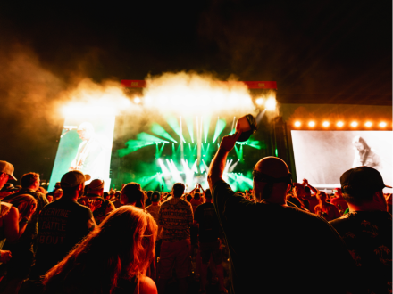 Crowd at an outdoor nighttime concert, arms raised, facing a brightly lit stage with green lights, smoke and giant screens.