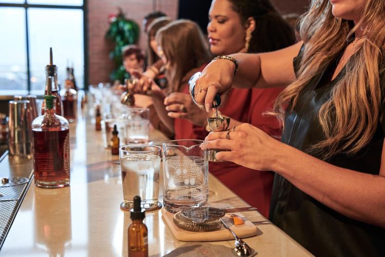 6 people standing at a bar and pouring alcohol into glasses during a cocktail class.