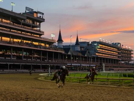 Pink and orange hues from the evening sky cast over a horse track with two racing horses at Churchill Downs.