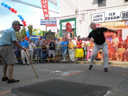 A man plays a sport in front of a restaurant with a stick and distance markers. A crowd cheers him on.
