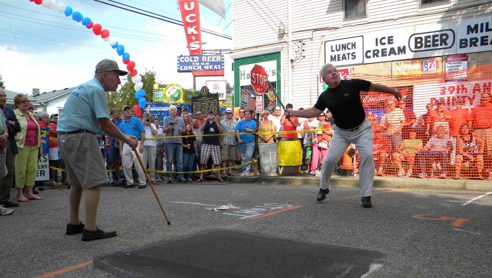 A man plays a sport in front of a restaurant with a stick and distance markers. A crowd cheers him on.
