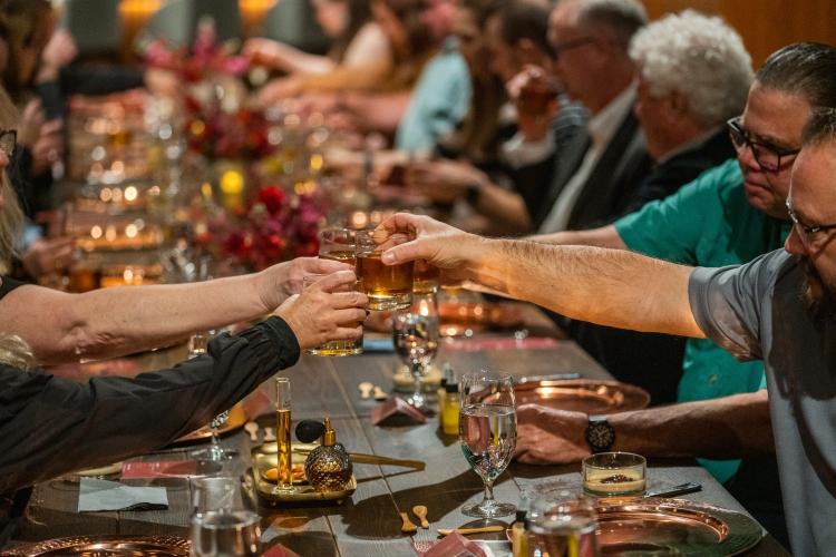 Four people extending their glasses out to cheers one another over a long candlelit table.
