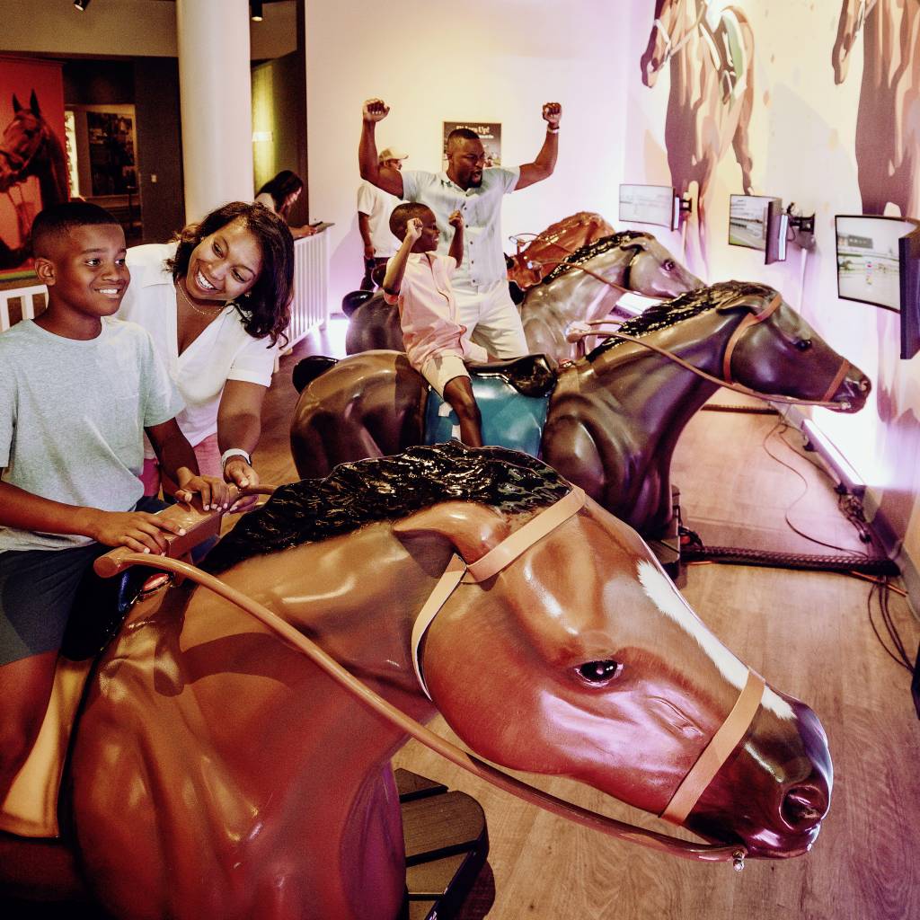 A family of four riding fake horses at an exhibit at the Kentucky Derby Museum.