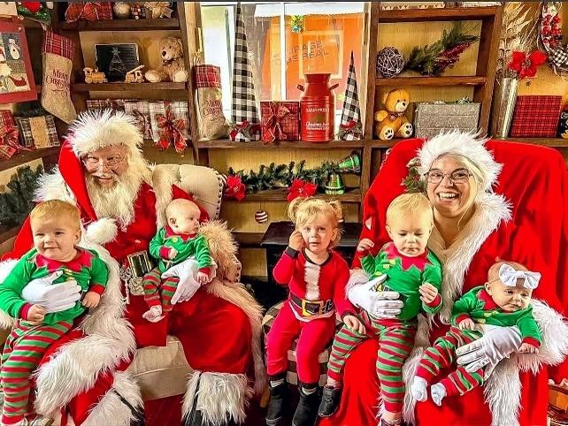 Santa and Mrs. Claus sit in chairs taking photos with 5 babies and toddlers dressed in elf costumes on their laps.