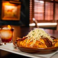 A Hot Brown sandwich in a baking dish on a plate piled high with bacon and cheese and an old fashioned cocktail.