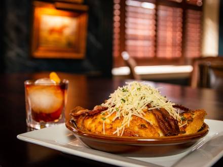A Hot Brown sandwich in a baking dish on a plate piled high with bacon and cheese and an old fashioned cocktail.