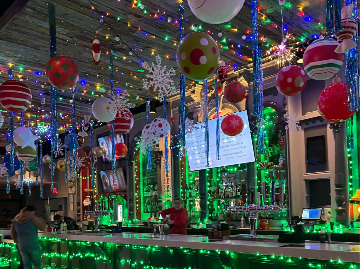 Giant green and red ornaments hang from the ceiling above the bar lined with green holiday lights at Tinsel Tavern.