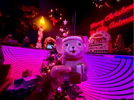Hand holding a  mouse-shaped holiday drink in a brightly lit Christmas pop-up bar with neon signs, wreaths and decorations.