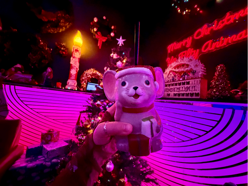 Hand holding a  mouse-shaped holiday drink in a brightly lit Christmas pop-up bar with neon signs, wreaths and decorations.