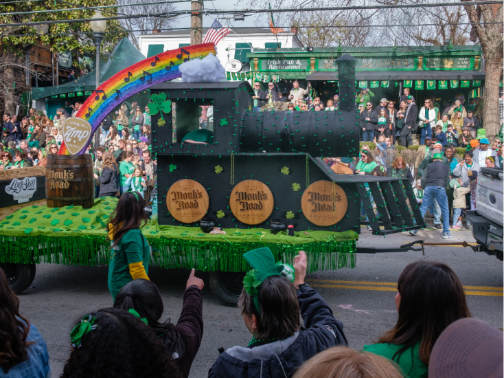 A green train with a rainbow shooting off the back sits atop a parade float as it moves down a street surrounded by a crowd.