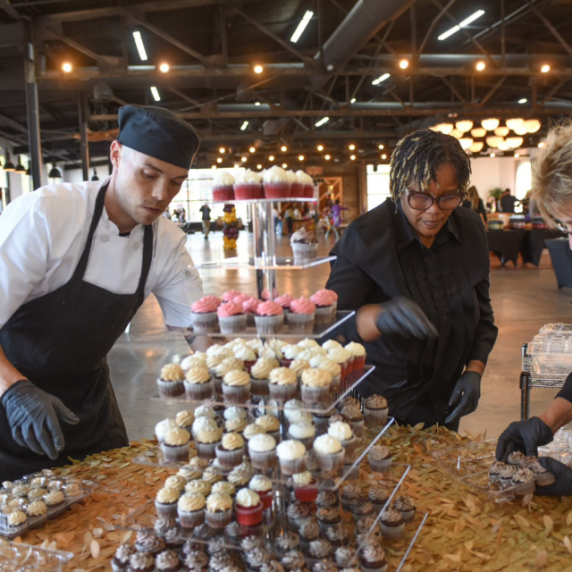 A three-tier dessert tray filled with mini cupcakes sits on a gold table with people surrounding.