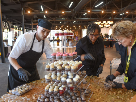 A three-tier dessert tray filled with mini cupcakes sits on a gold table with people surrounding.