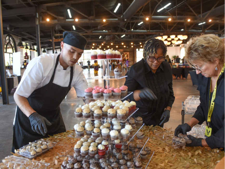 A three-tier dessert tray filled with mini cupcakes sits on a gold table with people surrounding.