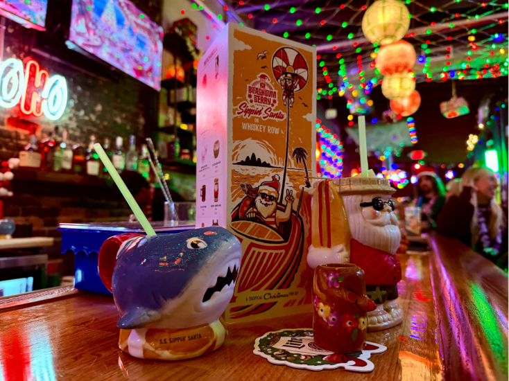 Festive holiday bar with Santa and shark tiki mugs on the counter, surrounded by colorful lights and cheerful décor.