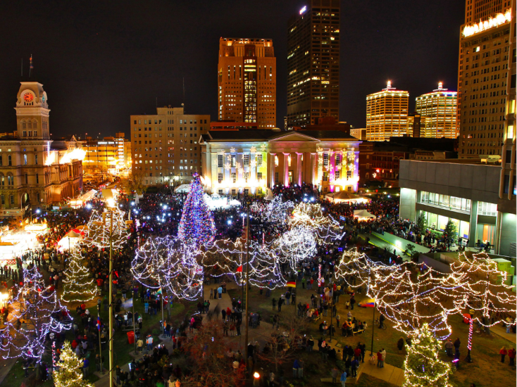 Crowds gather in downtown Louisville for a holiday celebration with lights, a Christmas tree, and festive skyline at night.