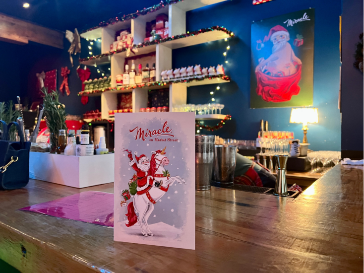 A holiday card sits on a wooden bar. Behind the bar are santa-shaped mugs and other holiday decor at Miracle on Market.