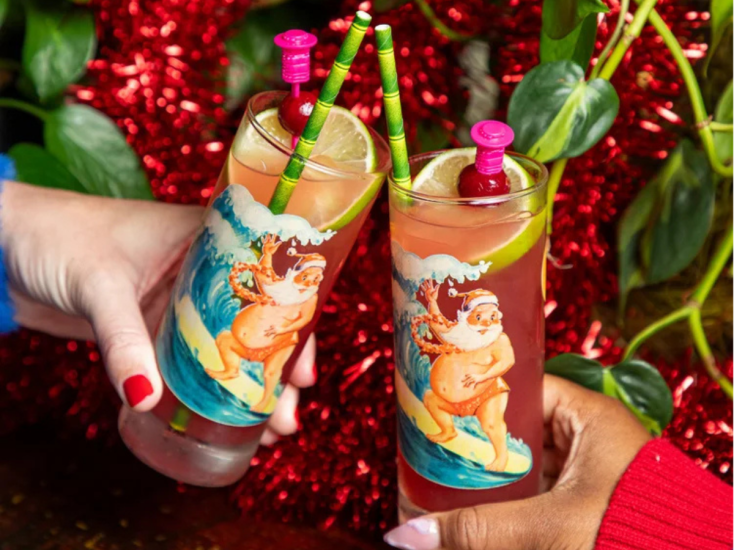 Two hands toast festive cocktails in surfing Santa glasses with lime and cranberry garnish against red tinsel backdrop