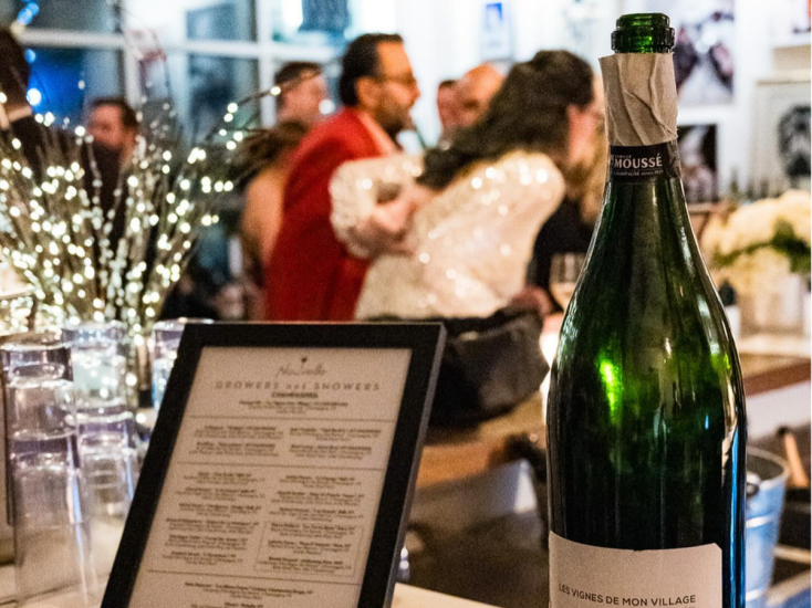 A champagne bottle and menu sit on a bar  at Nouvelle as guests mingle in a softly lit, festive setting.