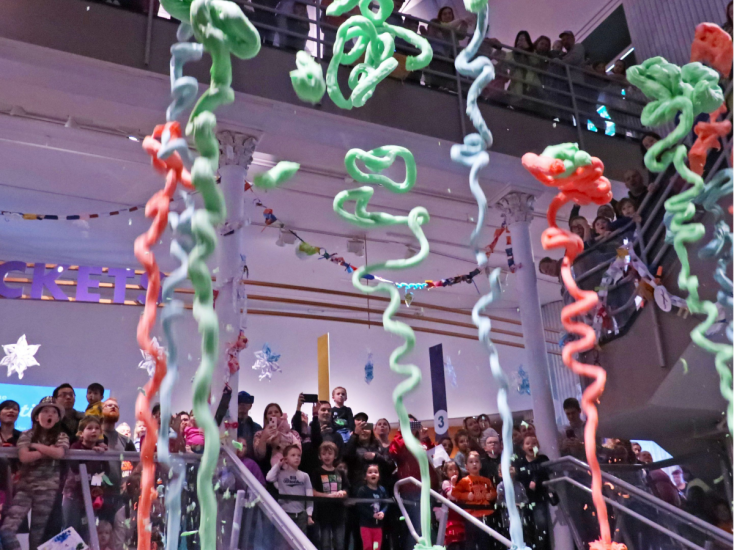 Crowd of kids and adults watch colorful foam streamers erupt indoors during a festive science-themed celebration.