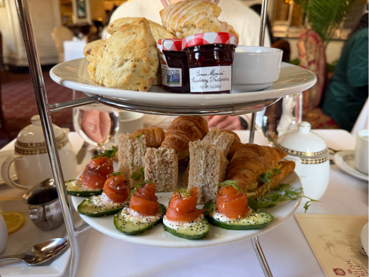 Tiered serving tray filled with scones, jarred jams, tea sandwiches, and cucumber slices.