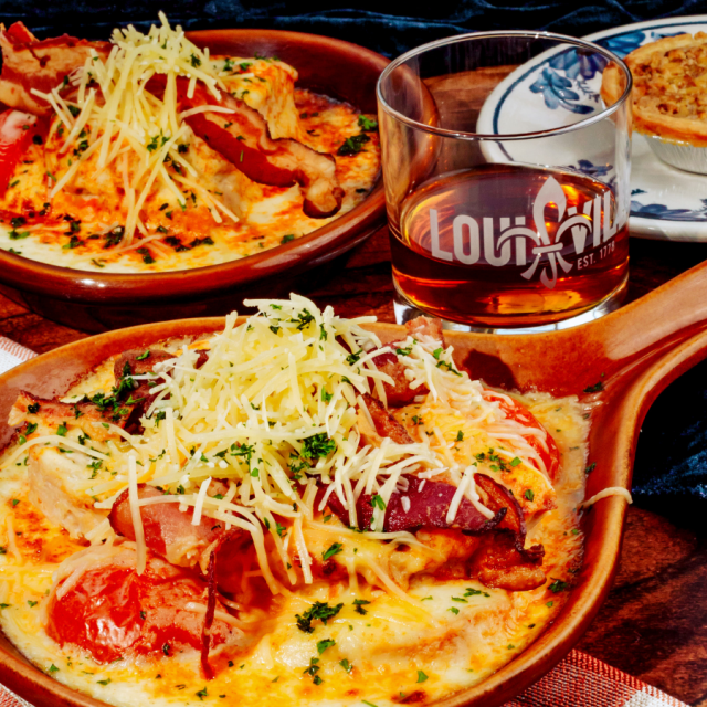Two brown crocks of baked turkey topped with bacon, tomatoes, and melted cheese beside a glass of bourbon and a small pie.