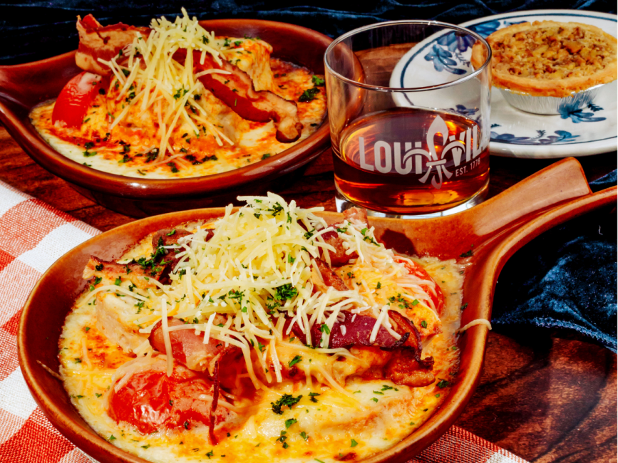 Two brown crocks of baked turkey topped with bacon, tomatoes, and melted cheese beside a glass of bourbon and a small pie.