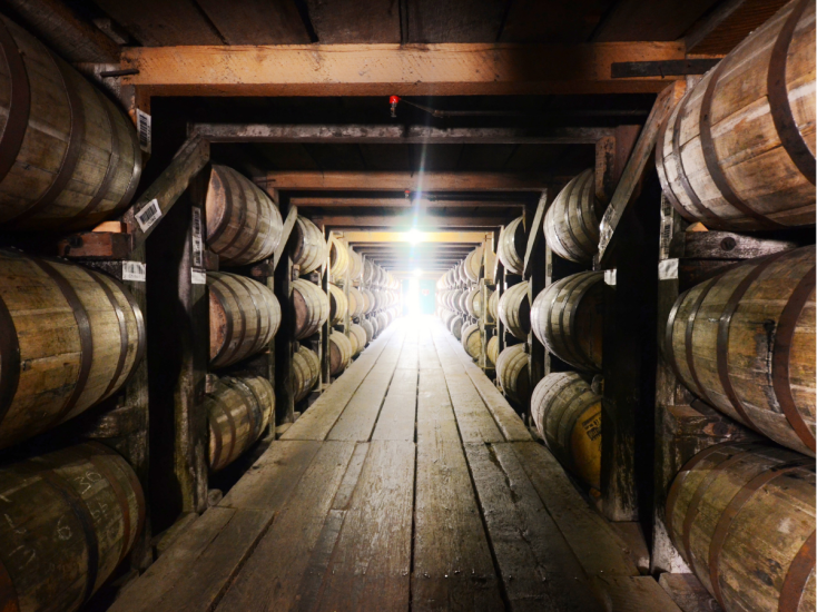 Light shines in through a door at the end of a wooden row of a rickhouse stacked with bourbon barrels three high.