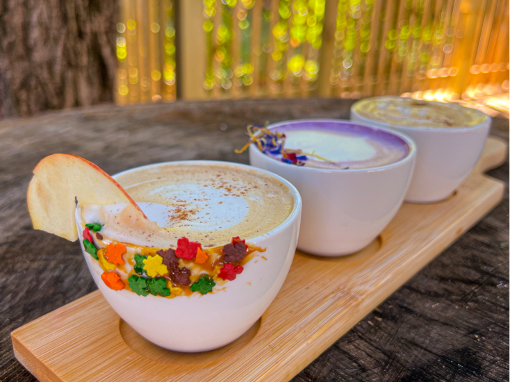 Three colorful phalcha coffee drinks served in white cups on a wooden tray, topped with festive garnishes and spices.