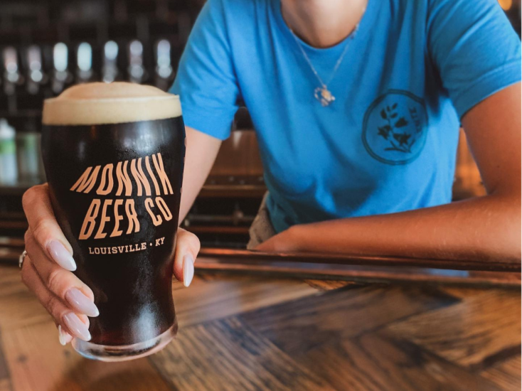 A person in a blue shirt holds a dark colored beer in a pint glass that reads "Monnik Beer Co." in orange letters.