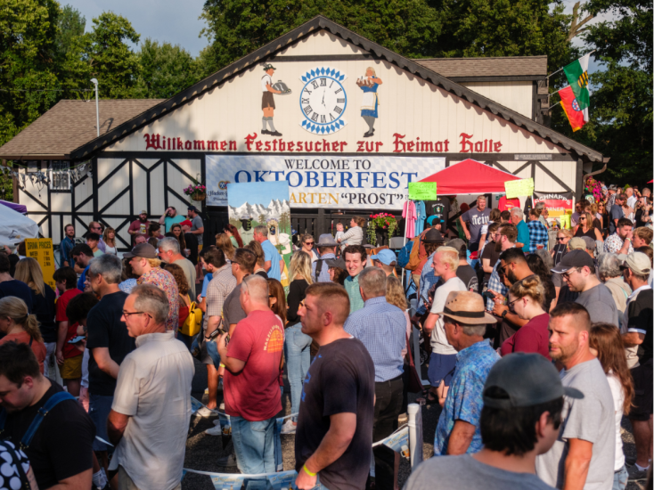 Crowds gather outside a festively decorated Oktoberfest hall with banners, signs, and people enjoying the lively celebration.