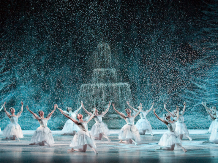 Louisville Ballet dancers in white costumes perform on stage, surrounded by falling snowflakes and wintry blue lighting.