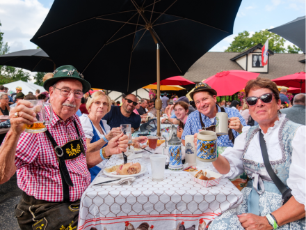 Group of people in traditional Bavarian attire raising steins and glasses while seated under umbrellas.