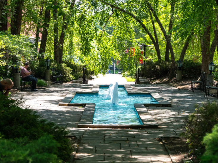 A shaded walkway with a cross-shaped fountain and benches surrounded by lush green trees at University of Louisville campus.