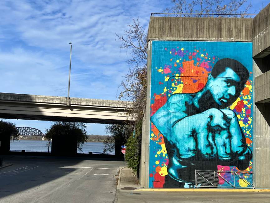 blue mural of Muhammad Ali on the side of a building near the water in Louisville