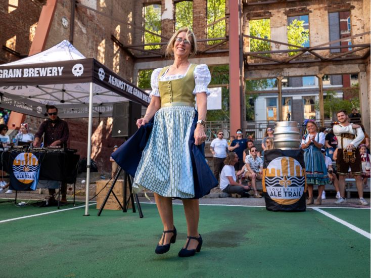 Woman in traditional dirndl dances at a Louisville Oktoberfest event, with music, beer kegs, and crowds in the background.