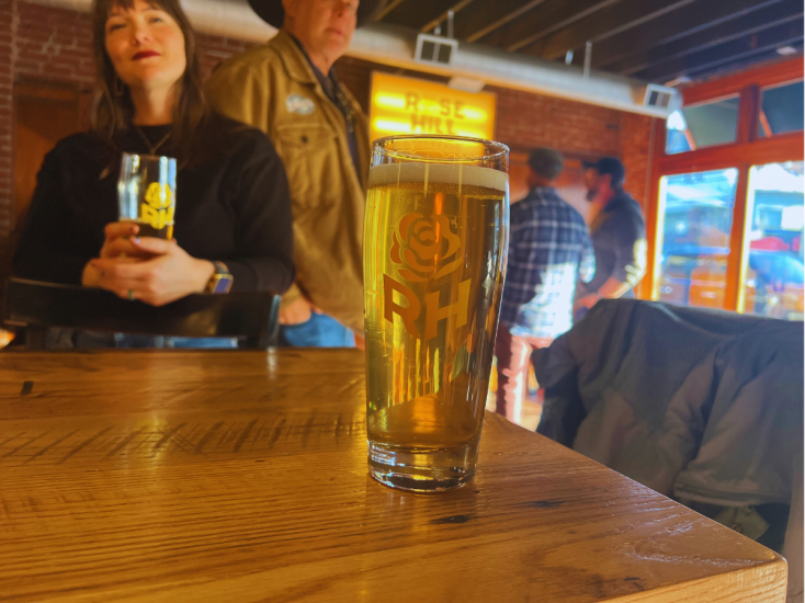 A pint of Rose Hill Lagerhaus beer on a wooden table, with patrons socializing inside the cozy Louisville brewery.