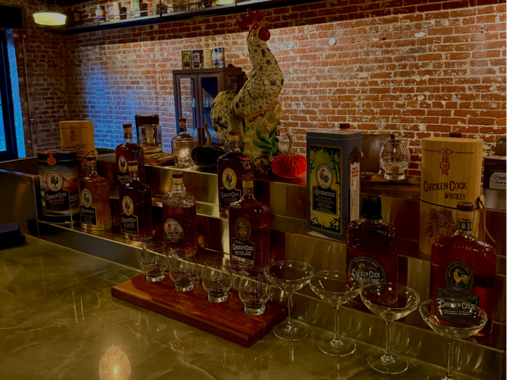 A bar displaying Chicken Cock Whiskey bottles, tasting glasses, and a ceramic rooster against an exposed brick wall backdrop.